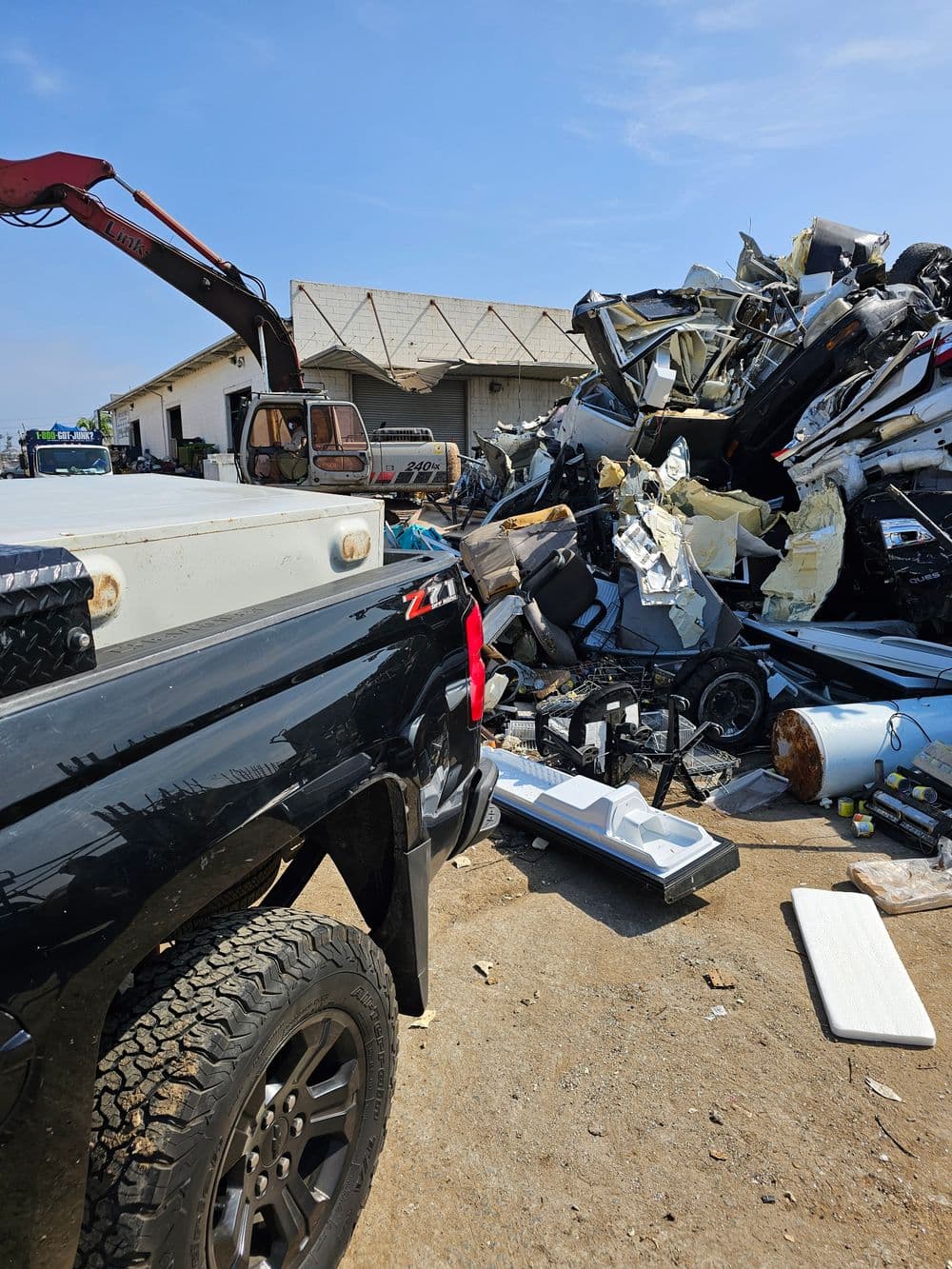 Black truck in foreground near a pile of crushed junk vehicles at a recycling yard.