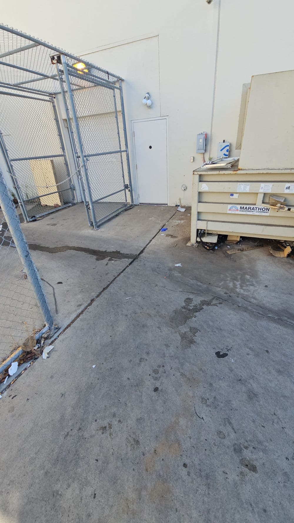 Metal dumpster near a fenced area and a small door, with a concrete surface showing stains.