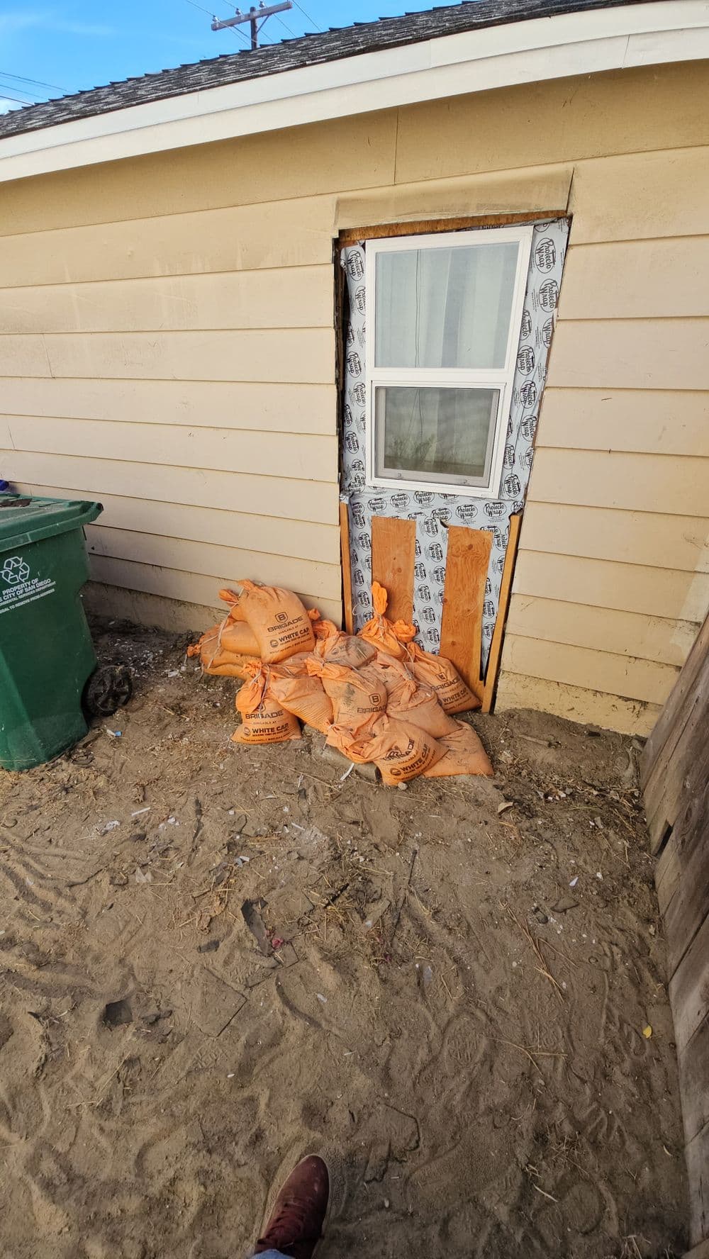 Sandbags piled against a door on a sandy surface near a green trash bin.