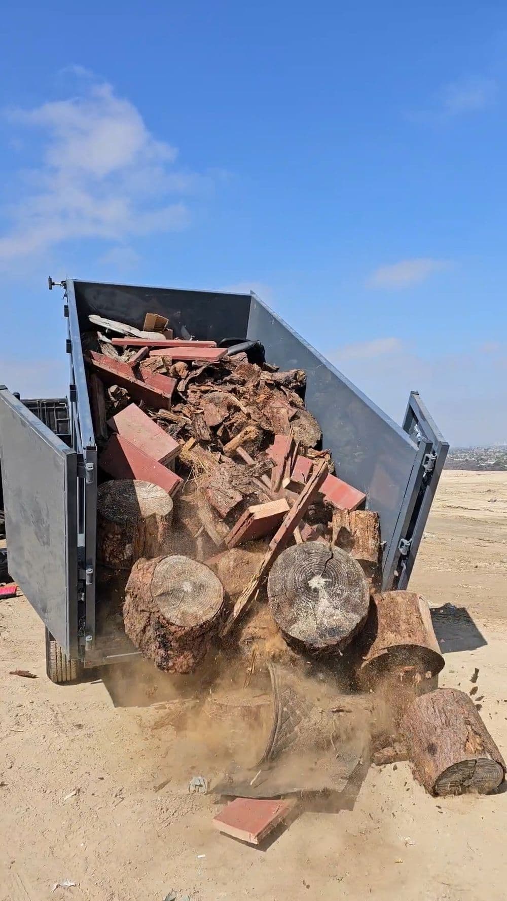 Dump truck unloading wood logs and debris on a construction site under a blue sky.