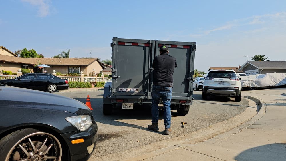 Man working on a gray trailer in a residential neighborhood with parked cars nearby.