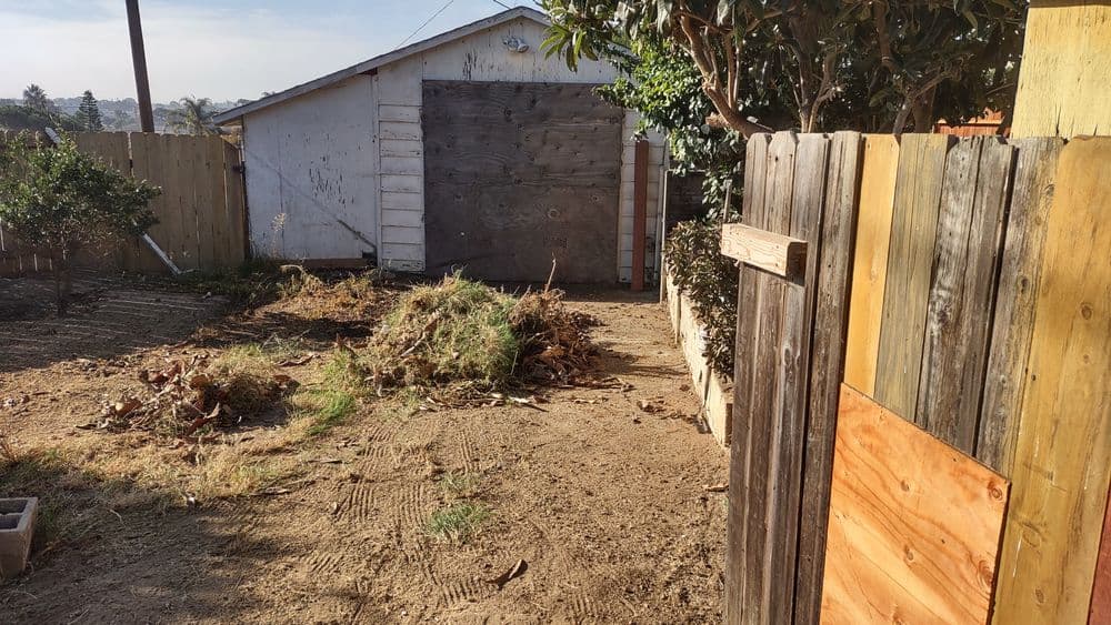 Overgrown yard with a wooden fence and a garage in the background, showing debris and dirt.